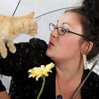 An image of Natalia, a person with dark hair, glasses, and a black shirt holds a yellow flower and looks at a small cat