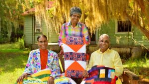 Three women hold brightly patterned quilts