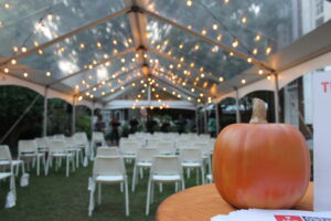 An orange pumpkin in front of rows of white chairs under a tent with fairy lights