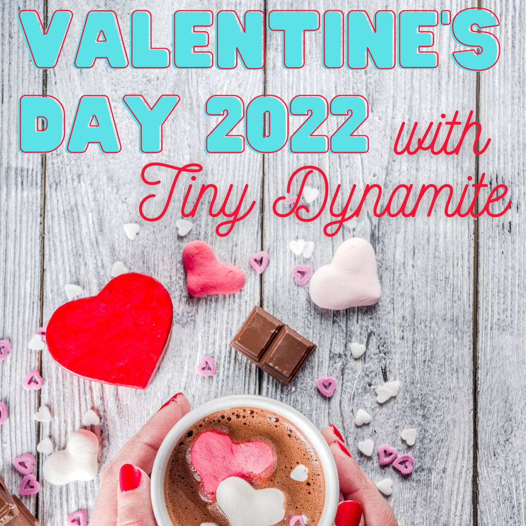 A hand with red nail polish holds a mug of hot chocolate with hearts and candy scattered around. The text says "Valentine's Day 2022 with Tiny Dynamite."