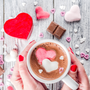 A hand with red nail polish holds a mug of hot chocolate with hearts and candy scattered around.