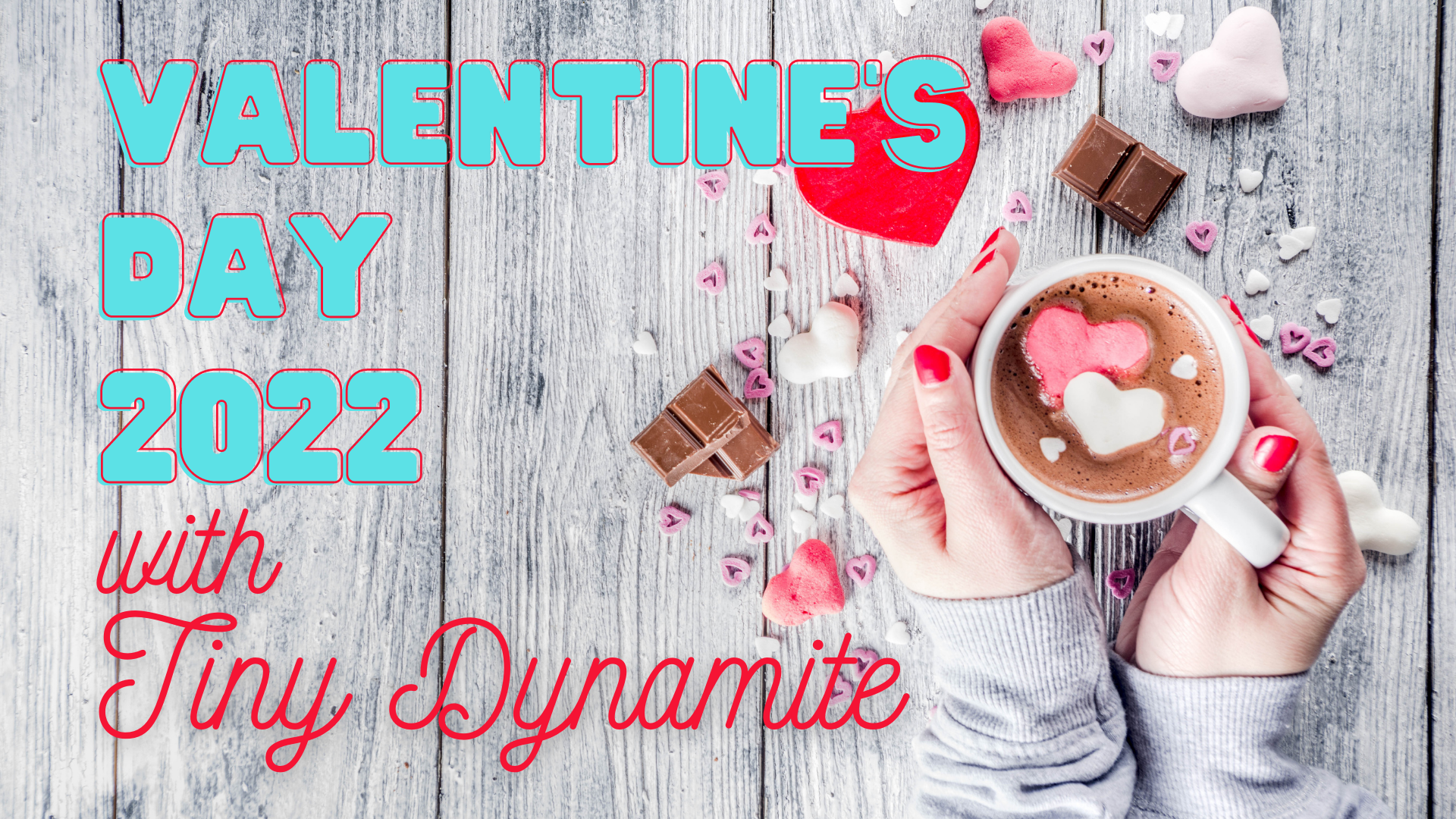 A hand with red nail polish holds a mug of hot chocolate with hearts and candy scattered around. The text says "Valentine's Day 2022 with Tiny Dynamite."