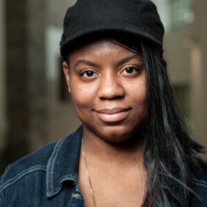 A woman with brown skin and black hair wears a black baseball cap and denim shirt and smiles at the camera.