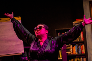 A performer wearing sunglasses standing in front of bookshelves stretches out their arms in purple lighting.