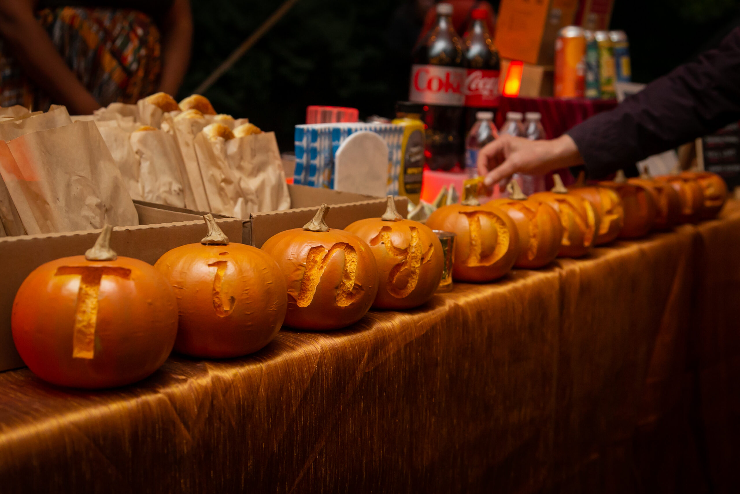 A row of small pumpkins with "Tiny Dynamite" carved into them, one letter at a time, in front of soft pretzels