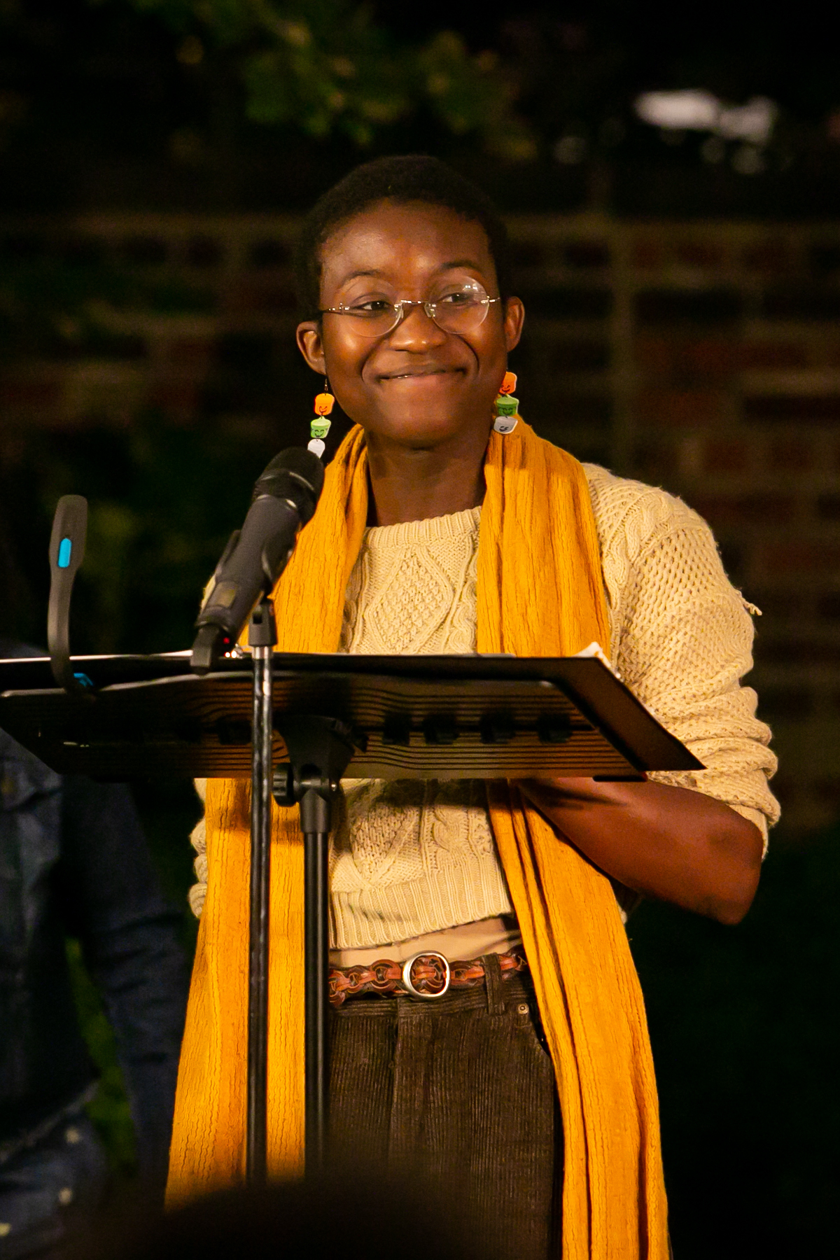 A performer in a yellow sweater and scarf stands adn smiles at a music stand
