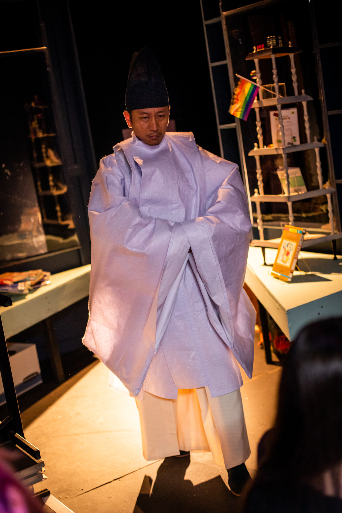 A performer in a white kimono and black hat enters, backlit with bookshelves behind him