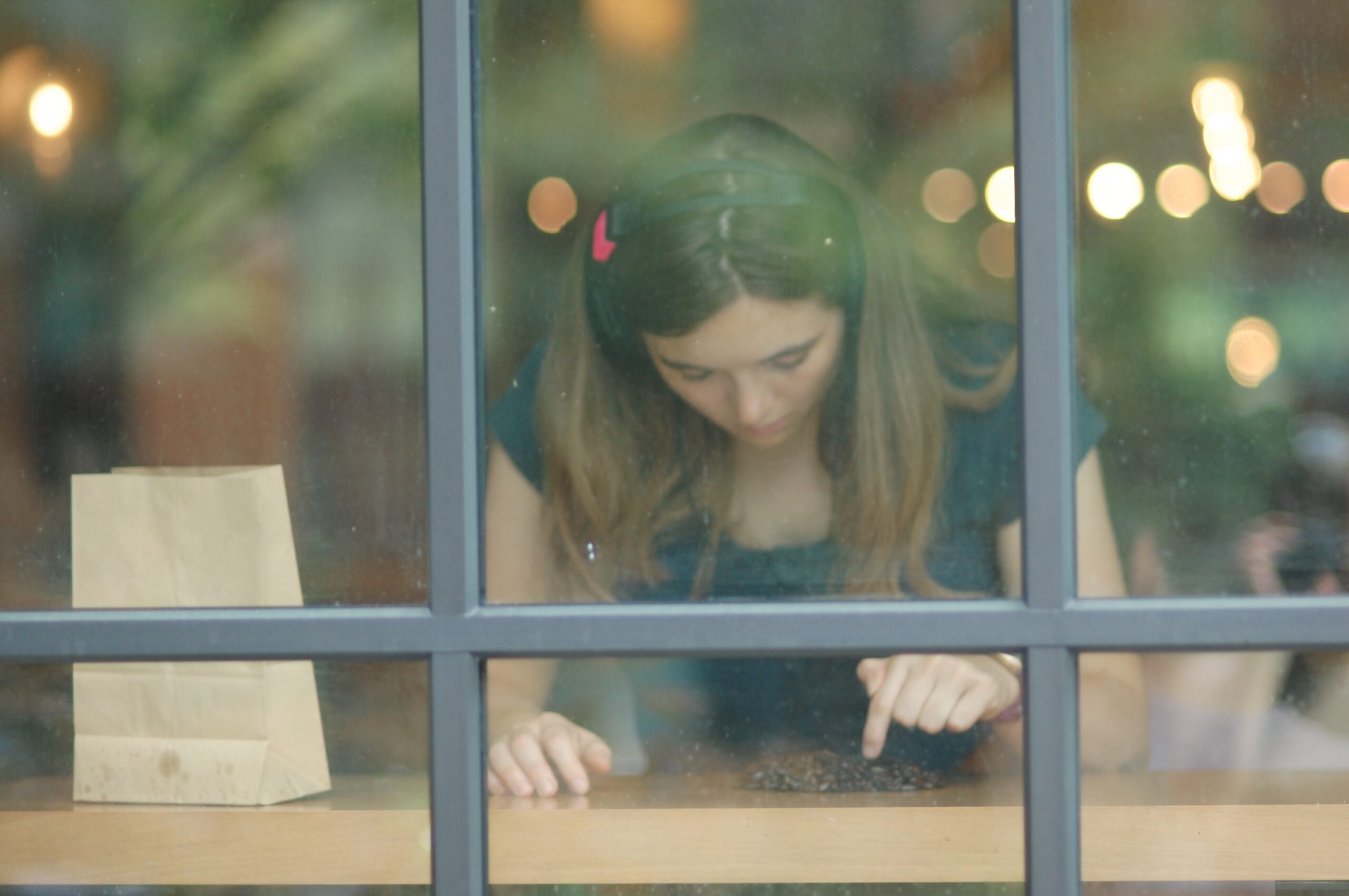 As seen through a window, a person sits at a cafe wearing headphones and counts coffee beans on the table